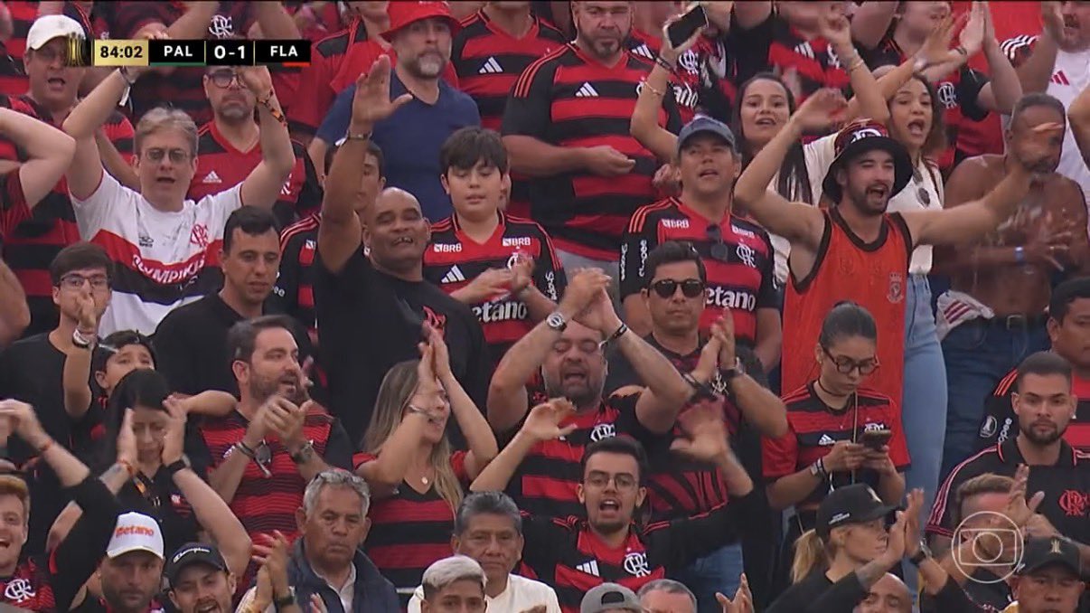 A imagem mostra um grande grupo de torcedores do Flamengo comemorando nas arquibancadas durante um jogo contra o Palmeiras, indicado pelo placar no canto superior esquerdo (84:02, PAL 0–1 FLA). A multidão veste majoritariamente camisas rubro-negras, algumas listradas, outras com modelos alternativos. As pessoas estão de pé, batendo palmas, levantando os braços e cantando, criando um clima de euforia. Há diversidade de idades: crianças, jovens e adultos. Um torcedor de camisa preta acena sorrindo no centro; outro, à direita, usa um chapéu bucket e uma regata laranja. Alguns olham para o campo, outros para celulares ou para a câmera. Ao fundo, veem-se mais pessoas em tons de vermelho, reforçando a atmosfera vibrante e lotada de um estádio em dia de jogo decisivo.