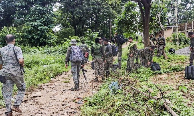 Group of uniformed security personnel in camouflage outfits and backpacks stand and kneel on a dirt path in a dense green forest with trees and vegetation around. Some carry rifles and inspect items on the ground including bags and a suitcase. The scene appears to be a post-operation inspection in a wooded area.