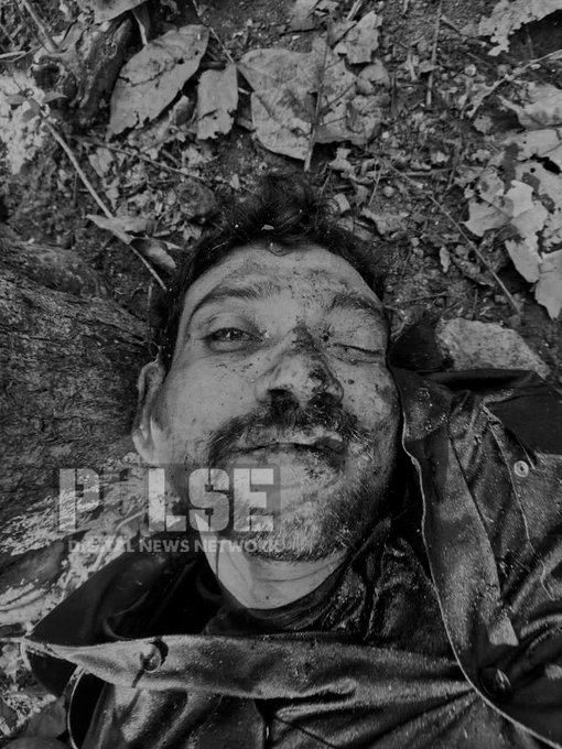 Black and white photograph of a man lying on his back on a forest floor covered with fallen leaves and dirt. His face and upper body are smeared with mud, eyes partially closed, mouth slightly open with a subtle smile, wearing a dark jacket. The scene appears post-struggle or operation in a wooded area.