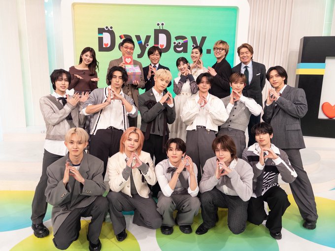 Group photo of 11 young male performers from INI standing and kneeling in formal suits and jackets on a brightly lit TV studio set with green and yellow DayDay logo backdrop heart symbol and floor markings some making hand hearts.