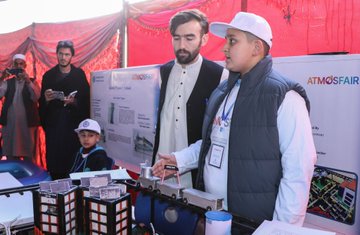 First image shows a group of men in traditional Afghan attire and suits standing on a stage holding award certificates for 25000 prizes with a backdrop featuring AtölyeSpark logo balloons and lights. Second image depicts men in traditional clothing and suits gathered around a table examining a green painted artwork with brushes and paints nearby. Third image captures individuals in masks and traditional outfits near a display of toy models including a hospital building train set solar panel and documents under a tent. Fourth image features people in traditional and modern clothing around a booth with toy building models of structures train and road setup plus a poster for AtölyeFair.