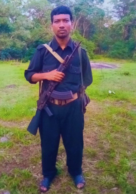 A man in a dark green uniform shirt and pants stands in a grassy forested area with trees and foliage in the background. He has short black hair, a mustache, and a serious expression. He holds an AK-47 style rifle slung over his shoulder with a strap across his chest. He wears sandals and carries a small bag on his side. The setting appears rural with dirt paths visible.
