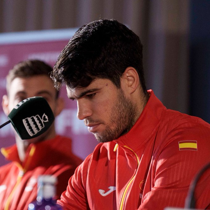 Carlos Alcaraz, a young male tennis player with short dark hair and a beard, sits at a press conference table wearing a red jacket with the Spanish flag and Joma logo. He looks serious and focused, facing slightly to the side. Microphones with logos are in front of him on the table, along with water bottles. Another person is partially visible in the background, and banners are on the wall behind.