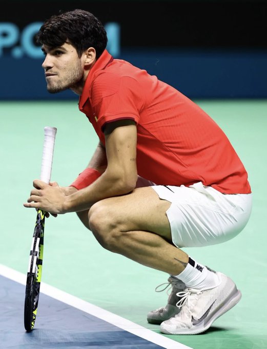 A male tennis player with dark curly hair kneels on a blue indoor tennis court surface near the baseline, wearing a red polo shirt, white shorts, white Nike sneakers with black accents, and holding a yellow and black tennis racket in his right hand. His expression is focused and serious, with a wristband on his right arm. The court lines and net are visible in the background.