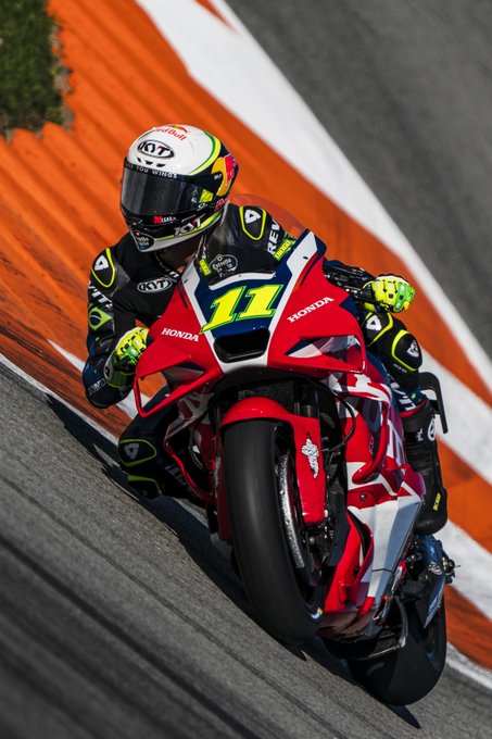 A professional motorcycle racer wearing a white helmet with red accents, black and green leathers, and yellow gloves rides a red Honda CBR1000RR-R motorcycle numbered 11 on a racetrack with orange curbs and white lines. The bike features LCR branding and is captured in a dynamic leaning turn. Green bushes and track barriers are visible in the background. The image is credited to Paco Díaz.