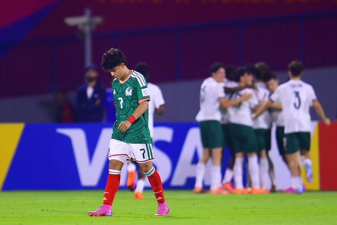 A dejected Mexican soccer player in green jersey with number 7 stands alone on the green field looking down wearing red wristband and pink cleats while in background a group of Portuguese players in red jerseys numbered 3 and others celebrate hugging near the sideline with yellow and purple stadium seating and VISA sponsor banner visible on the advertising board behind them under stadium lights.