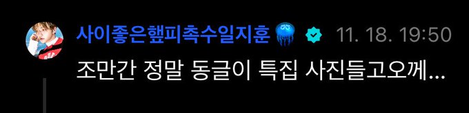 A young man with long dark hair sits on a bed in a simply furnished bedroom. He wears a black tank top and looks directly at the camera with a neutral expression. The background includes a large window with sheer curtains, a green plant on the windowsill, and beige walls.