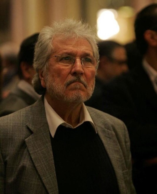 Elderly man with gray curly hair, glasses, mustache, and beard wears a gray suit jacket, white shirt, and black sweater. He stands in a warmly lit interior space with blurred background of other suited men.
