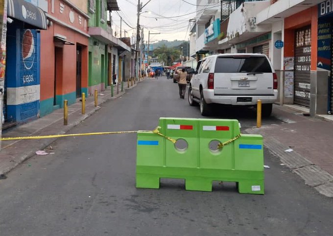 A street scene in an urban area with colorful buildings painted in red green and blue along both sides lined with shops and power lines overhead. A white SUV is parked facing the camera in the middle of the road. Yellow caution tape stretches across the street blocking access with green plastic barriers placed at intervals. People stand in the background near a red bus and more vehicles. The setting appears to be in a hilly neighborhood during daytime.