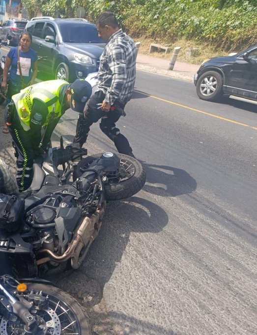 Roadside scene in a green hilly area with several cars parked including a black SUV and silver sedan. A man in plaid shirt stands near the vehicles. Two individuals in high-visibility yellow-green vests and black pants inspect a fallen black motorcycle lying on its side with visible exhaust and wheels exposed. Another motorcycle stands upright nearby. Bystanders including a woman in blue shirt observe from the side.