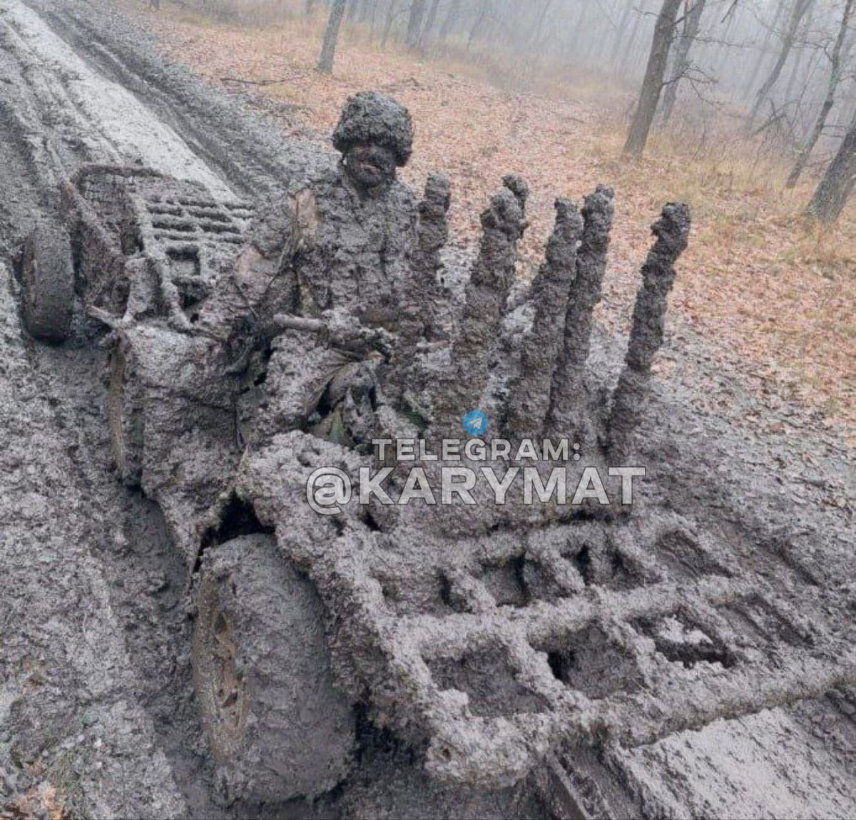 Soldier wearing camouflage helmet and uniform sits on a four-wheeled ATV vehicle covered in thick mud, steering along a narrow dirt road surrounded by bare trees and fallen leaves in an autumn forest setting. The ATV has a cargo rack loaded with several long muddy poles or branches stacked upright. Foggy atmosphere pervades the scene with distant trees visible. Watermark from @karykarmatmat appears on the image.