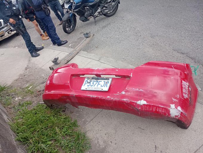Red cars rear bumper detached and lying on the street with visible dents and scratches, displaying license plate P537FJR. Three police officers in dark uniforms and boots stand nearby observing. A black motorcycle is parked close to the bumper. A white truck is visible in the background on the road. Roadside grass and a wooden plank are present beside the scene.