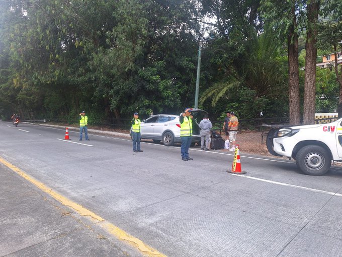 Roadside scene on a paved road with green trees and palms in background several workers wearing yellow high-visibility vests and helmets stand near a white sedan and a white truck with Provial markings orange traffic cones placed around the area a motorcycle parked nearby and a man in civilian clothes with luggage beside the vehicles.