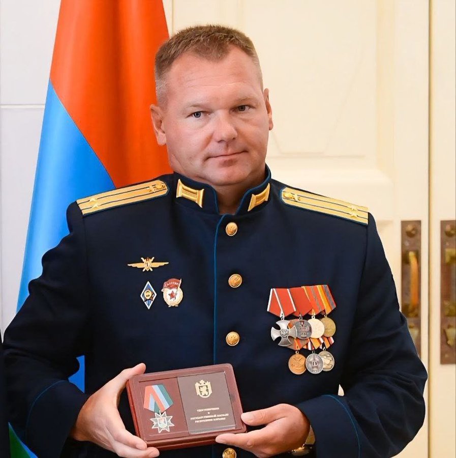 一名身穿深蓝色军服、佩戴金色肩章、胸前佩戴多枚勋章和胸针的男子站在一扇木门前，门两侧是俄国旗帜和另一面国旗，手中握着一块带有两个徽章的木质牌匾。