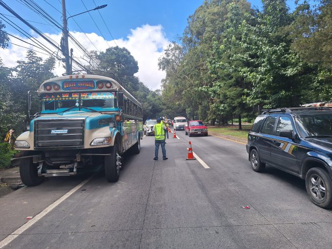 A blue and white interurban bus with decorative elements is stopped on the right side of a paved road under a clear sky with scattered clouds and green trees in the background. A worker in a high-visibility yellow vest stands in the middle of the road directing traffic with orange traffic cones placed around the area. Several other vehicles including a red car and a dark SUV are visible on the road nearby with power lines overhead.
