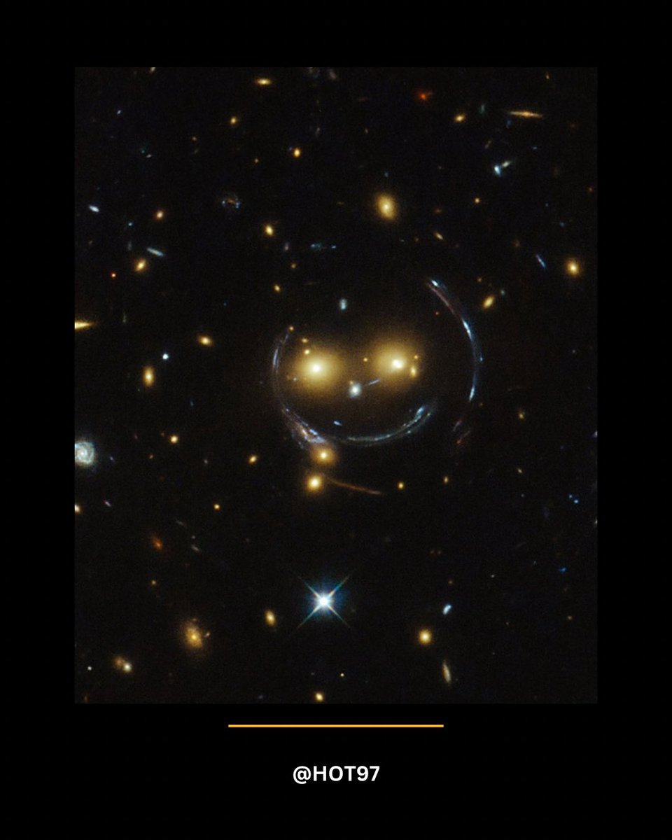 HOT97's tweet image. Hubble snapped a wild “cosmic smiley face” made from two glowing orange galaxies forming the “eyes,” a faint white glow for the “nose,” and curved arcs shaped by intense gravitational lensing that create the “smile.” These galaxy clusters are so massive they distort spacetime…