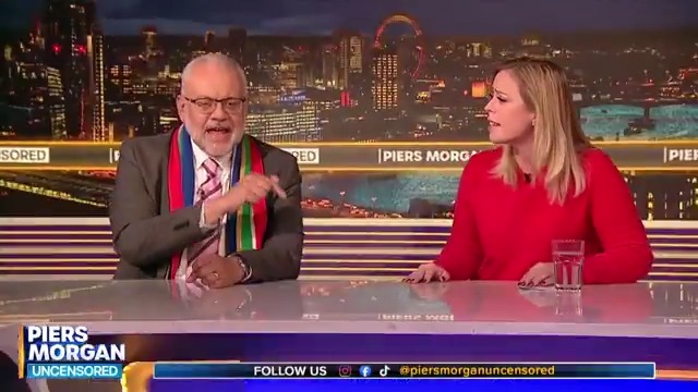 A split-screen studio interview on the Piers Morgan Uncensored show at night, featuring a male guest in a dark suit with a colorful sash (resembling South African flag colors: red, white, blue, green, yellow, black) gesturing while speaking, and a female guest in a red sweater listening attentively. The background shows a nighttime London skyline with the Thames River and city lights; the desk has water glasses and Piers Morgan logo overlays.