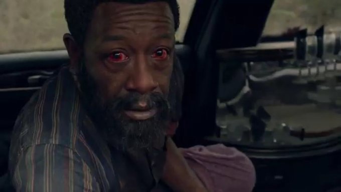 A close-up portrait of an African American man with a beard, wearing a striped shirt, sitting inside a dimly lit car interior; he has intense red glowing eyes, an open mouth expression of surprise or intensity, and is looking directly at the viewer; background includes car dashboard elements and a blurred window.