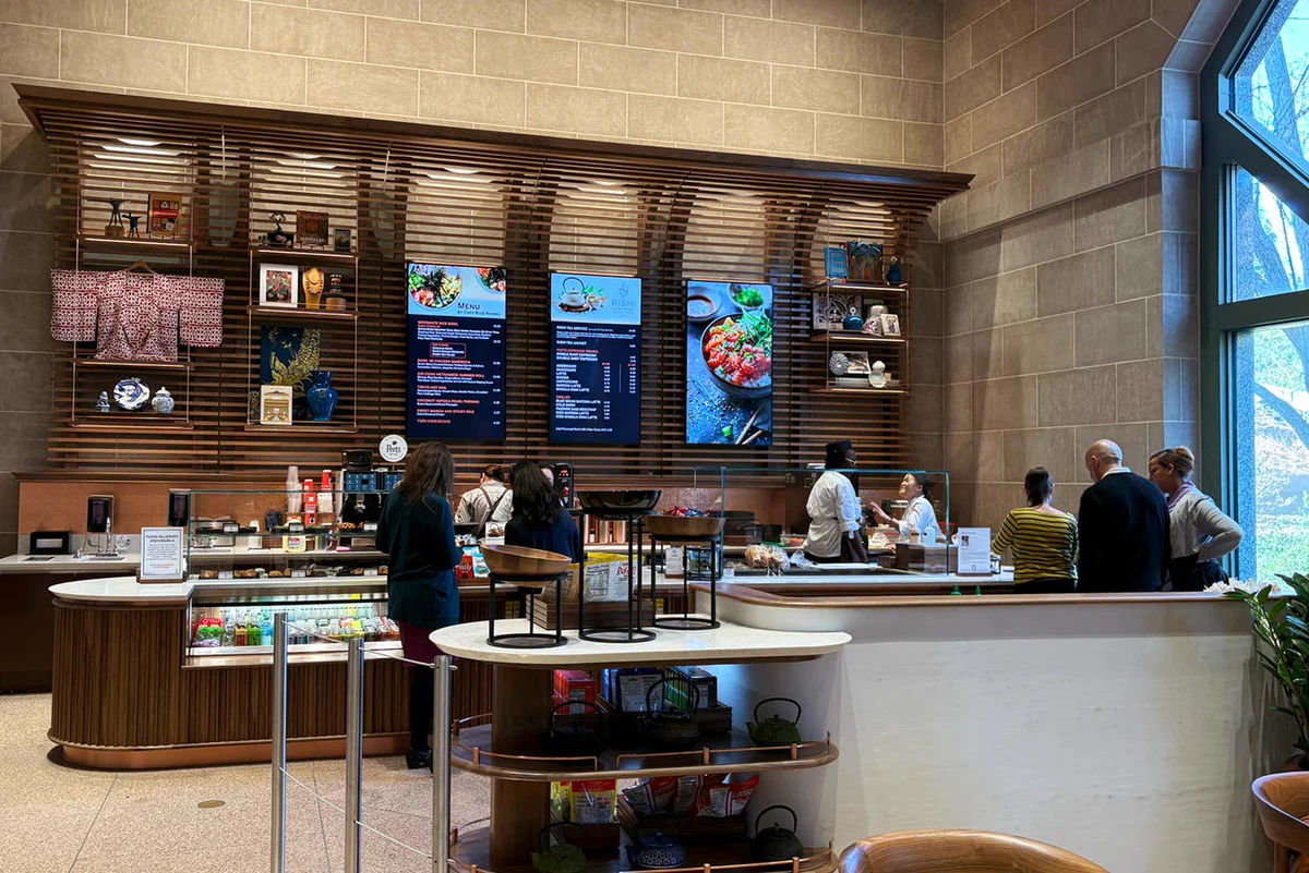 Interior view of the Moongate Cafe at the National Museum of Asian Art, showing customers ordering at the counter and others seated at tables. The background features digital menu boards above the counter.