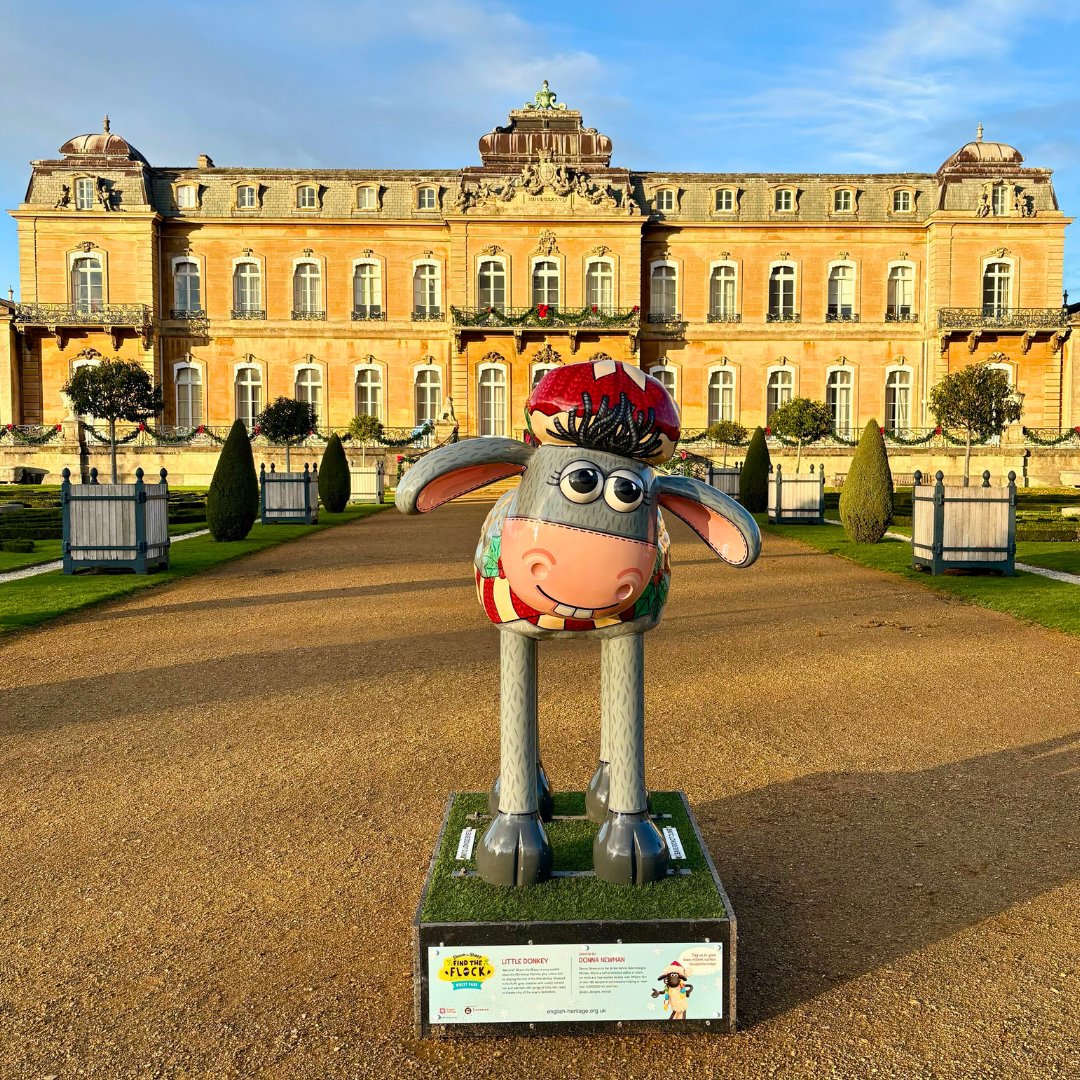 A colourful sheep sculpture is dressed up as a donkey and is wearing a winter hat and sweater. The statue stands in front of a grand, ornate building under a blue sky.