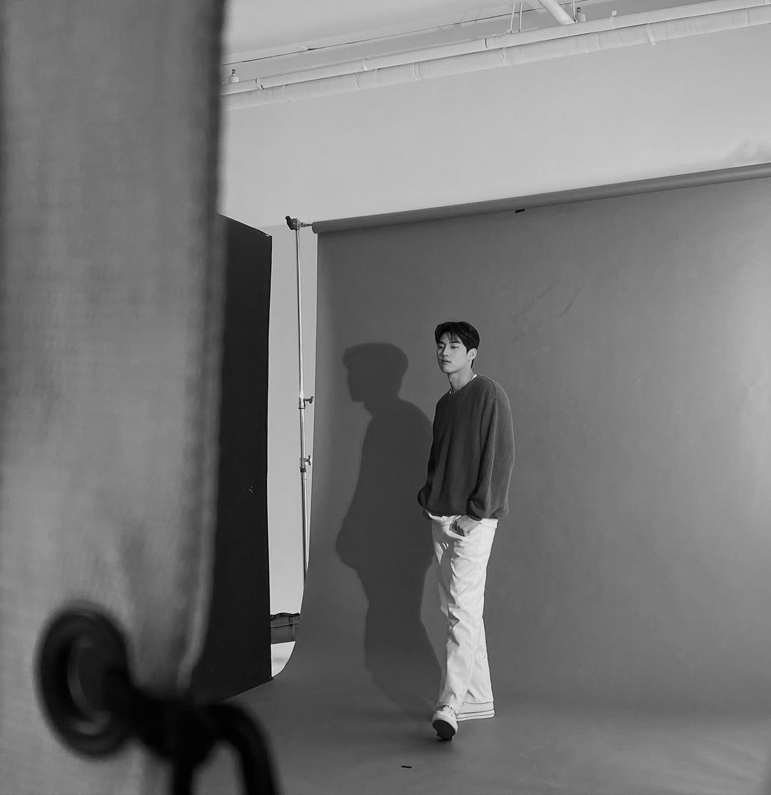 Dozyun's tweet image. He stood in the studio with a calm, effortless confidence, his relaxed posture enhancing the clean and minimal atmosphere around him. The monochrome setting highlighted the soft textures of his sweater and the crisp look of his white pants.