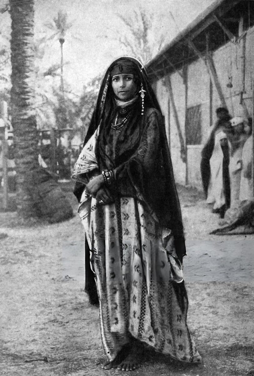 IRQMso's tweet photo. سيّدة شيعية من عرب الأهوار، تقف بكامل أناقتها تحت ظِل النّخلة؛ رأسها مغطى بـ”العَصّابة أو الچرْغَد“ – الزيّ التقليدي في الأهوار وأرياف الجنوب – وهو وشاح مصنوع من القماش الناعم الذي تتخلّله خيوط جميلة من البريسم، كما ترتدي حُليًّا منها أقراط طويلة متدلّية وقلائد، إضافةً إلى أساور في معصميْها. ألتُقطت في مطلع عام ١٩٠٠مـ. محفوظة في الأرشيفات: [Fireshot Studio/Fototeca/Universal Images Group].