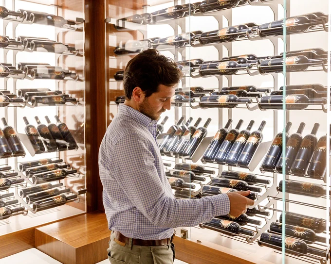Tenuta Luce’s enologist standing in the Collection room, holding a wine bottle with one hand. The display consists of multiple rows of horizontally arranged wine bottles on sleek metal racks inside a glass enclosure. The setting has warm wooden accents and bright lighting that highlights the bottles.