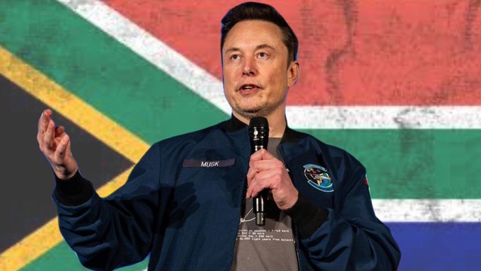 The image depicts Elon Musk standing at a podium, speaking into a microphone while gesturing with his hands. He is wearing a dark blue jacket with a 'MUSK' name tag and a SpaceX patch. In the background, the South African flag is prominently displayed.