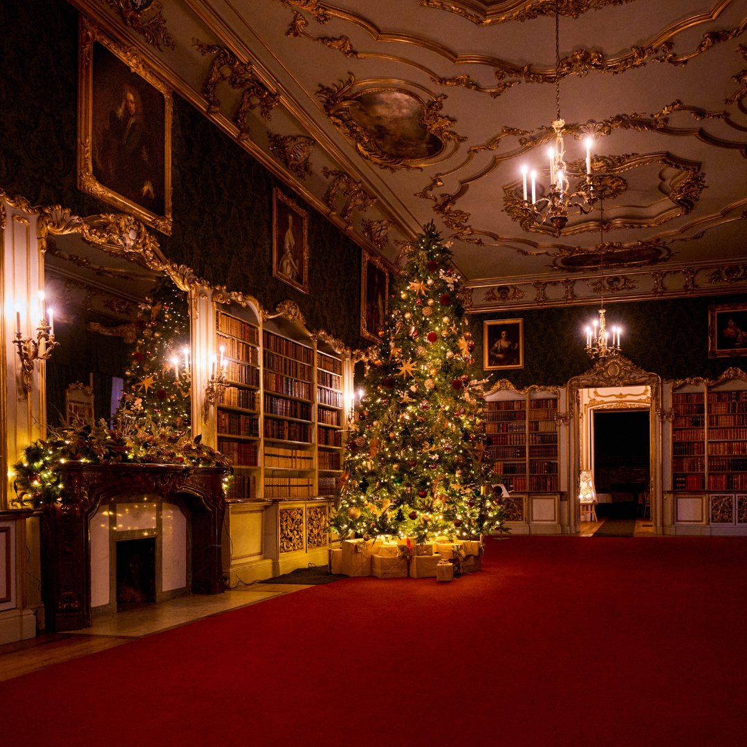 Luxurious room with ornate ceiling, golden-framed paintings, and a grand Christmas tree adorned with glowing lights and presents, evoking warmth and elegance.