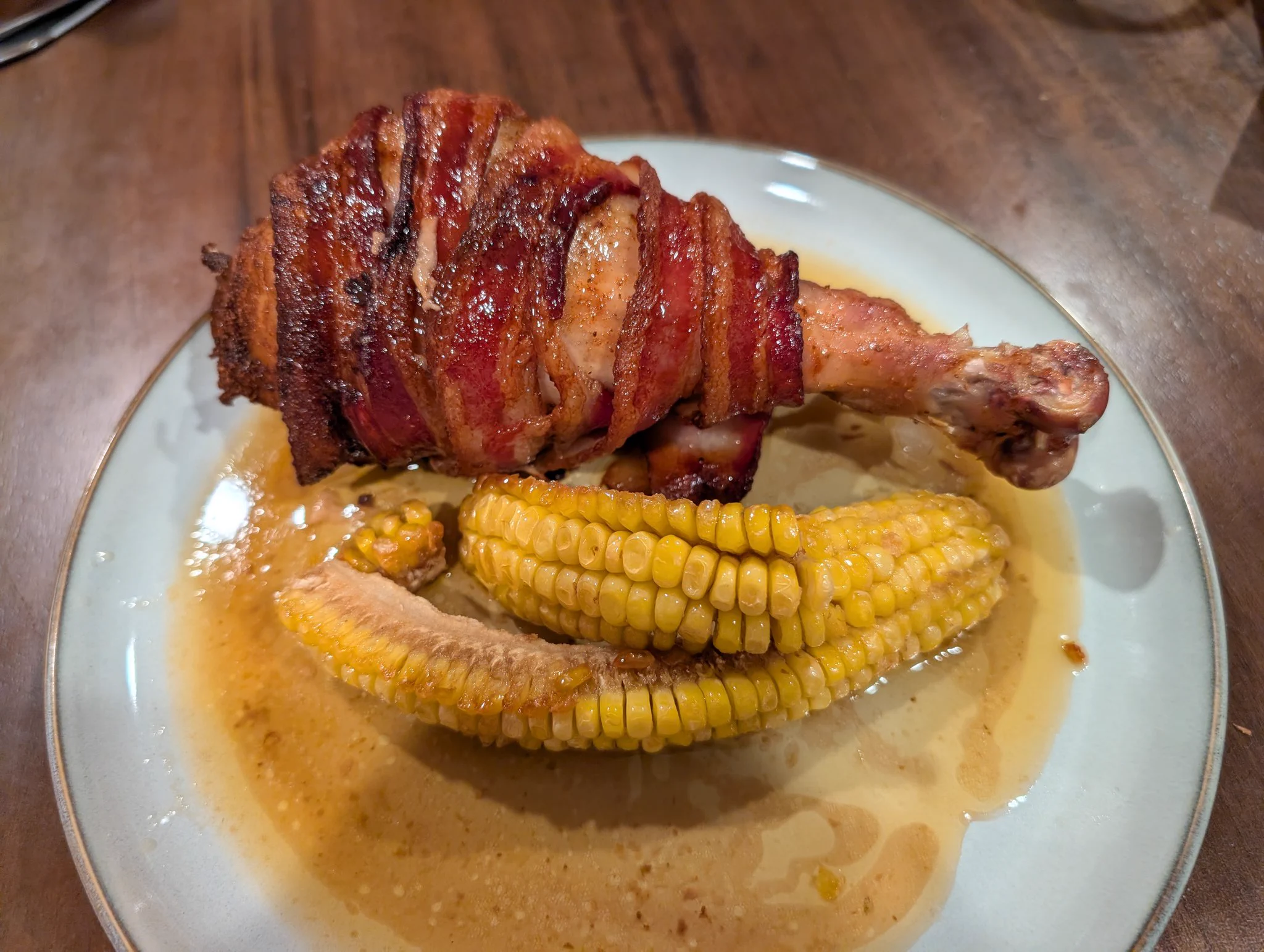 A dinner plate with a large bacon-wrapped turkey leg and some corn ribs. 
