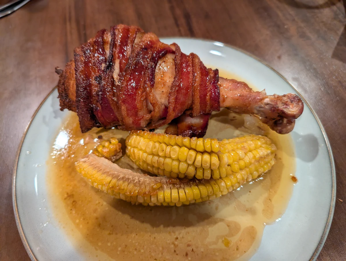 A dinner plate with a large bacon-wrapped turkey leg and some corn ribs. 