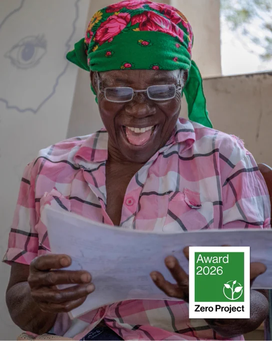 A woman wearing glasses, a green headscarf, and a pink checked shirt smiles brightly while reading a sheet of paper. She sits indoors with soft natural light. A green “Zero Project Award 2026” badge appears in the lower right corner of the image.