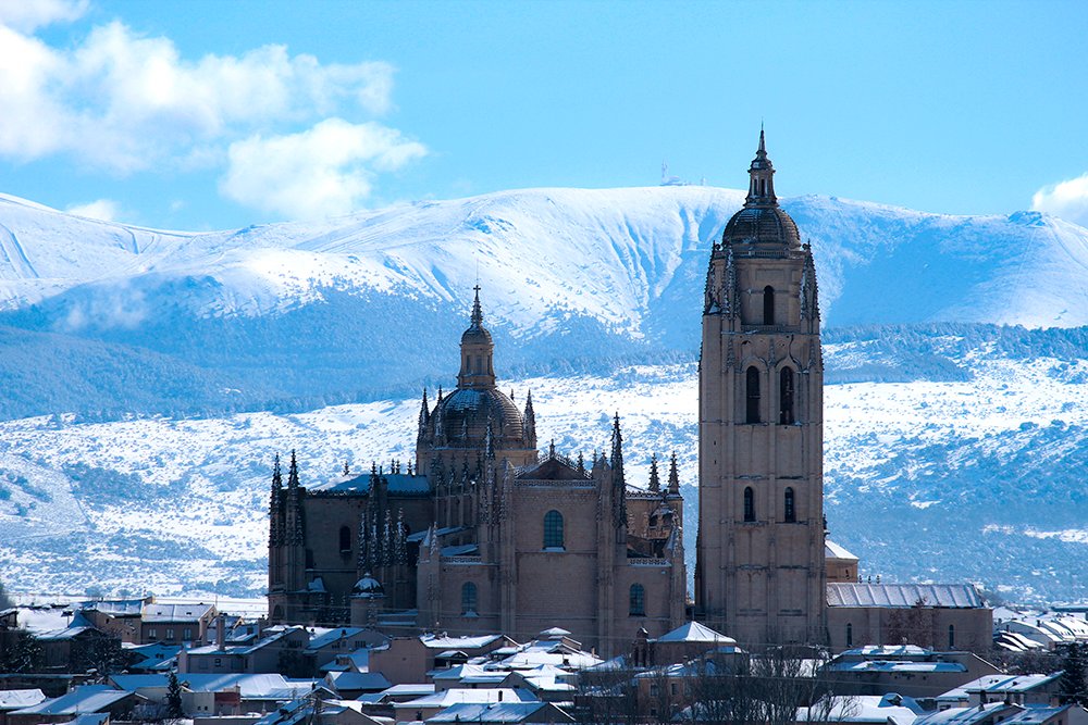 La Dama de las Catedrales, resurgiendo de la nieve. Su silueta gótica se recorta contra un cielo frío y limpio. Coronando todo, la vertiente segoviana de la sierra de Guadarrama, con la Bola del Mundo brillando en lo alto. segoviaunbuenplan.com