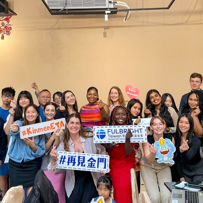 Group of people smiling and holding signs with hashtags and names including "#KimmelETA" and "Fulbright." Decorations hanging in the background suggest a festive or celebratory event.