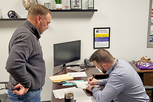 Hampton High School Principal Carson Klute signs paperwork Monday night to become the next Hampton Public School superintendent next summer when Holly Herzberg retires.  At left is school board president Grant Dose. See this week's ANR for all the details.
