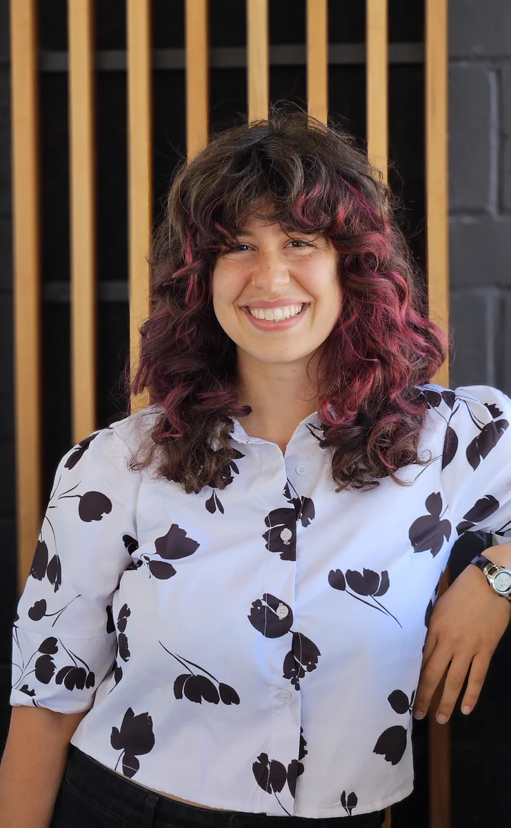 A headshot of Amanda smiling in front of a wood panelled wall. Amanda has long dark curly hair with pink highlights, light skin, and is wearing a white blouse with black flowers on it.