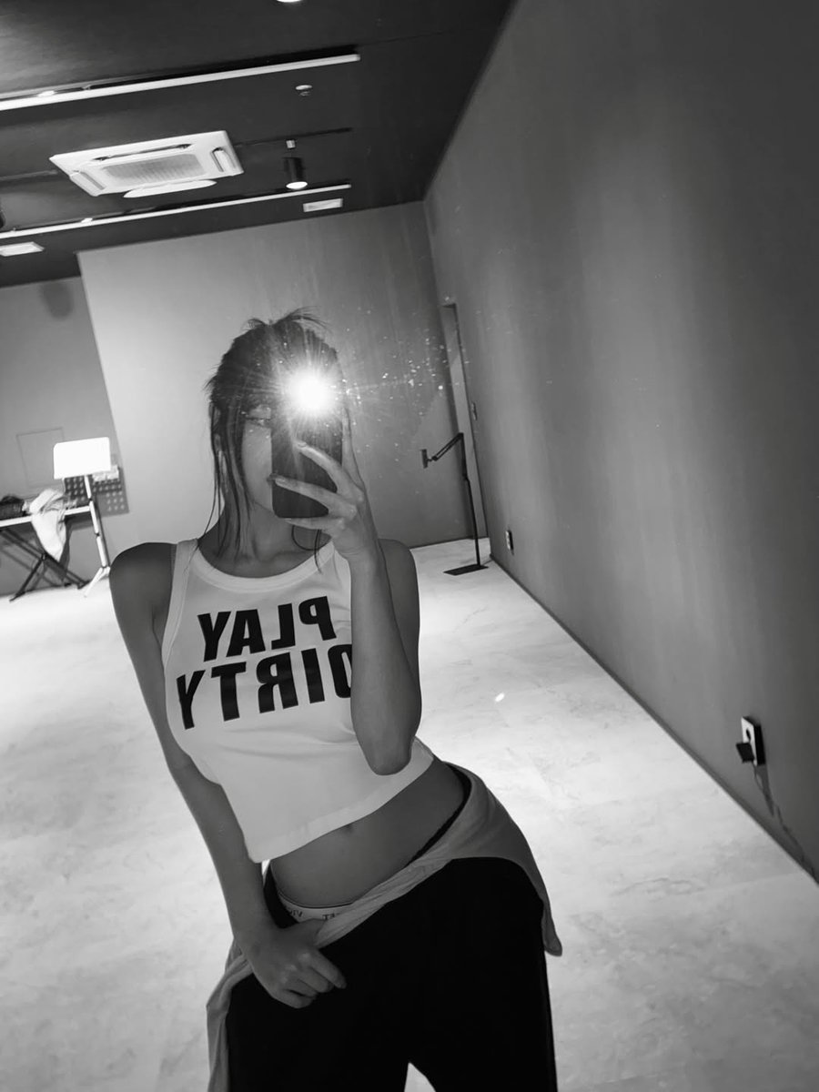 Flash on, vibes immortalized in black and white! That raw mirror moment — cropped white tank with the bold upside-down statement 'PLAY DIRTY' hitting different, midriff out, messy hair framing the mystery, and the phone flash exploding like a supernova right in the center.