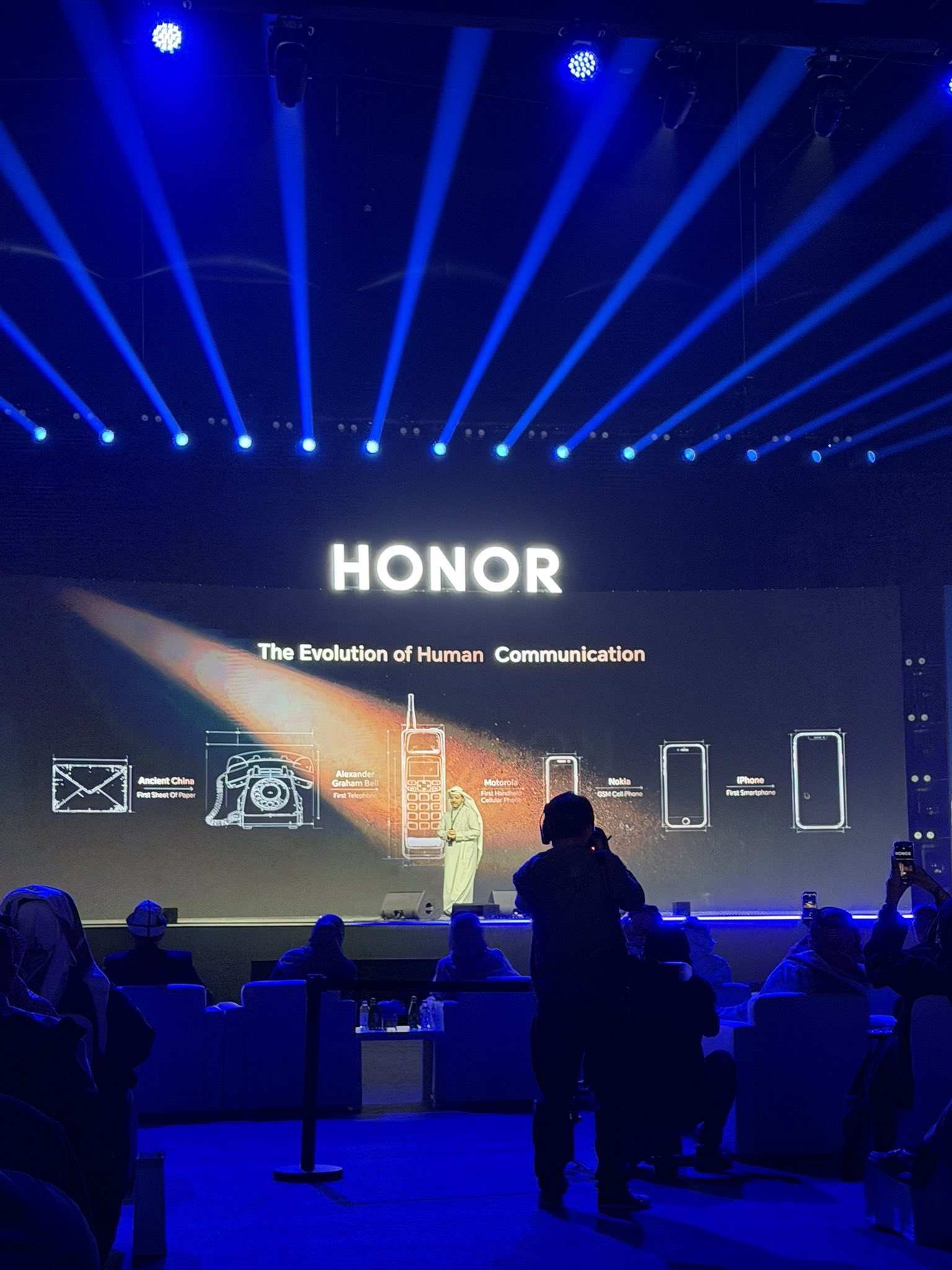 النص الظاهر في الصورة وترجمته: HONOR. تطوّر التواصل البشري. من بدايات التواصل في الصين القديمة. إلى اختراع أول هاتف على يد ألكسندر غراهام بيل. ثم أول هاتف محمول من موتورولا. وأول هاتف GSM من نوكيا. وصولًا إلى الآيفون كأول هاتف ذكي #سحر_بلمسة_واحدة 