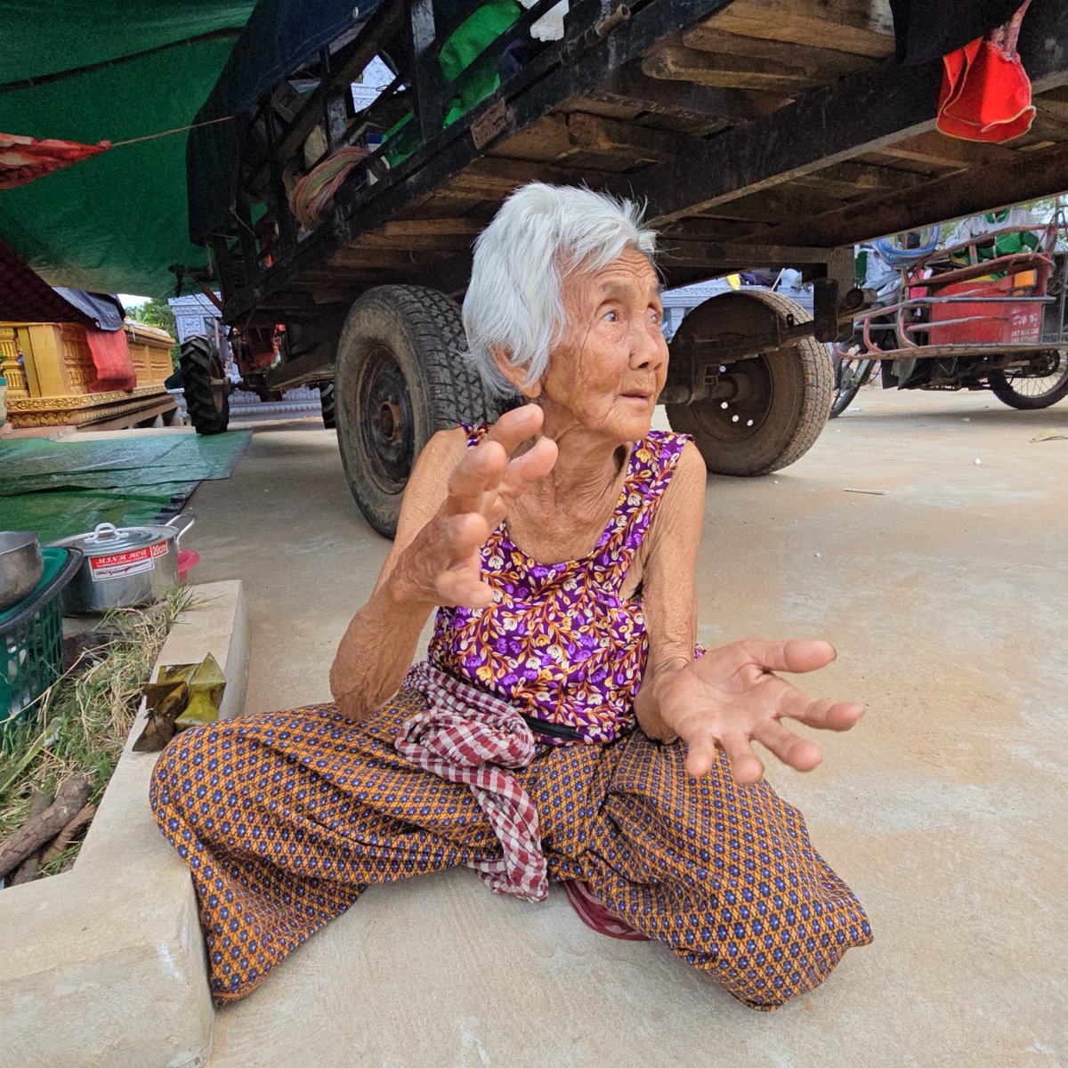 TTNEnglish's tweet photo. “I Could Not Sleep All Night”: A 93-Year-Old Woman’s Fear in a Displacement Center

BANTEAY MEANCHEY – Por Long, 93, is among the few families forced to evacuate from Svay Chek commune in Svay Chek district, Banteay Meanchey province, following intensified Thai military bombardment.
Her family fled on December 11 after repeated explosions and the presence of military drones made it impossible to remain in the village.
“We could not stay because many bombs were falling and so many military drones,” Long said.

Long has nine children, but none of her biological children live with her due to long-standing financial hardship. She lives alone and receives care from her daughter-in-law, who resides nearby.

She is currently staying at Kralanh Pagoda displacement center in Siem Reap province with several hundred other families.

Long said she fled with her daughter-in-law and one grandchild on a homemade tractor, bringing very few belongings.
“I came here, and I could not sleep at night. I am afraid, afraid of Thai fighter jets,” she said.

“Last night I sat awake the whole night. When I heard an explosion, I kept waking my daughter and grandchild to run for safety.”

The ongoing violence has revived memories of Cambodia’s civil war. Long recalled her father's advice: during wartime, travel against the flow of water to avoid danger.

“It might be superstition, but it is something that has protected me since I was young,” she said. “I always followed his advice to stay safe.”

Like many elderly evacuees, Long struggles with loneliness after being displaced from her home. Her greatest wish is to return as soon as possible.

“I want to go back home. I miss it so much,” she said. “I keep urging my daughter to drive back, but our village is still being bombed.”

The bombardment has not stopped, and Svay Chek continues to be heavily targeted by Thai artillery and shelling. Although Long longs to return, she fears the fighting may not end soon.

“I am not sure because the bombs are in their hands,” she said. “If they want to attack, they can keep doing it even if we urge them to stop. We cannot do anything else.”

“Maybe we have to stay here until the bombs fall silent.”
Despite her age, Long continues to tend a small garden at home, growing produce to sell so she does not have to rely on her children, who live far away. 

Some send her small amounts of money, but it is not enough to cover her daily needs.

“I left three rows of vegetables at home when we fled,” she said. “We were among the few families who stayed even when the shelling became intense.”

“I do not know if my vegetables are still good. Maybe they have already been damaged.”

Photos by Sao Phal Niseiy