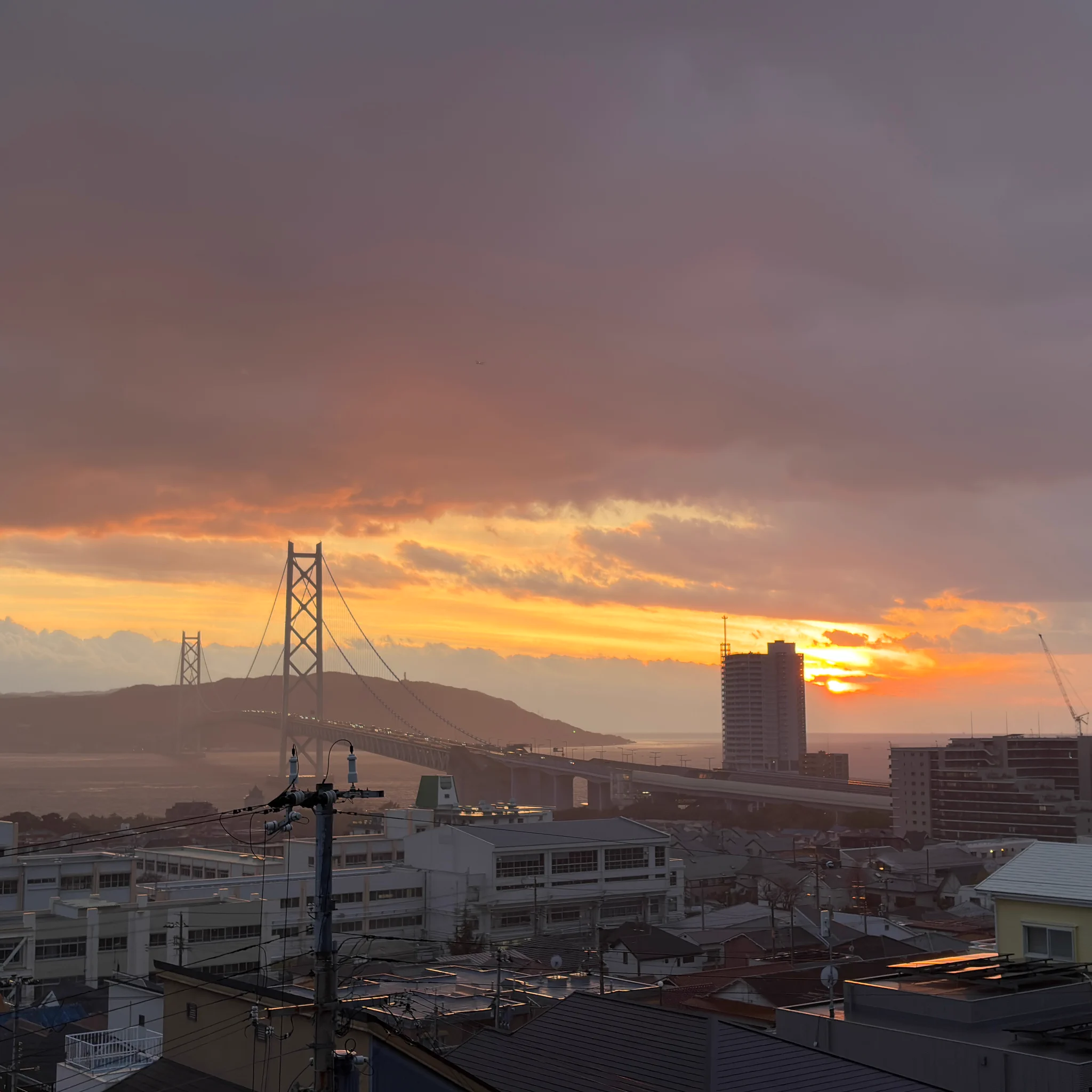 Sunset over a coastal Japanese city with orange light under dark clouds; bridge and buildings in the distance
