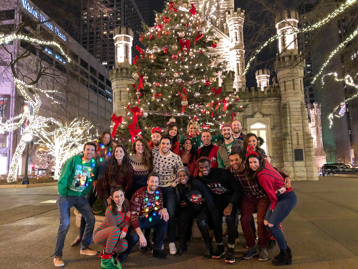 Group of people posing in front of a large, beautifully decorated Christmas tree at night, surrounded by illuminated trees, near Chicago's iconic Water Tower Place.