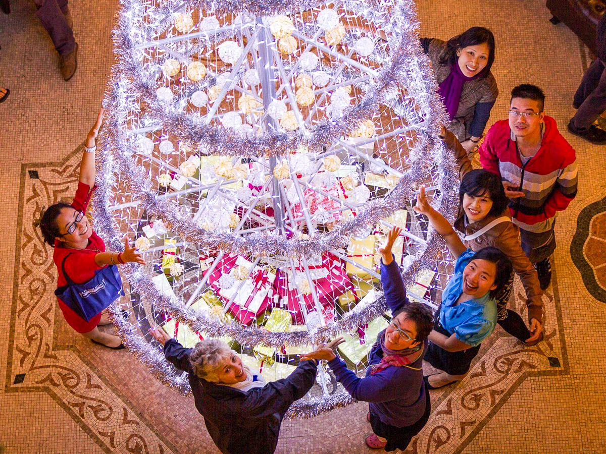 Group of people joyfully reaching towards a large, decorated Christmas tree from below, viewed from an overhead perspective.