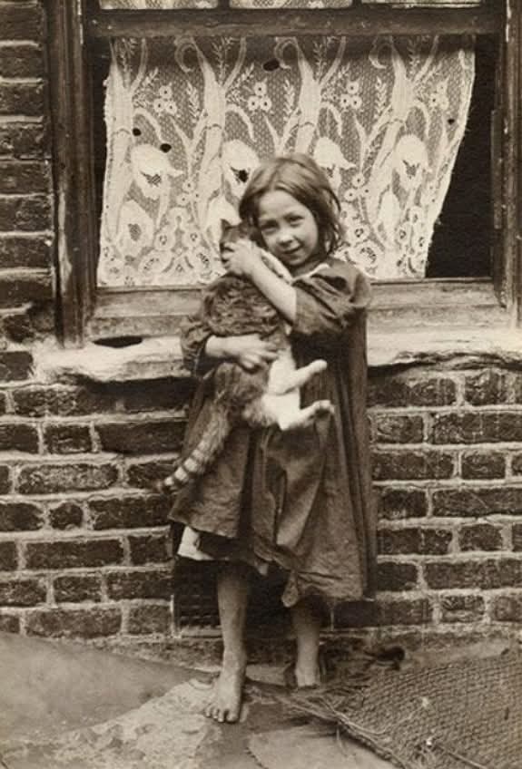 Dr_TheHistories's tweet image. In this quietly powerful photograph from 1902, Horace Warner captures a barefoot young girl seated on a stoop in London’s East End, gently holding a cat close to her chest. Her clothes are worn, her expression solemn, and the setting stark—but the way she cradles the animal…