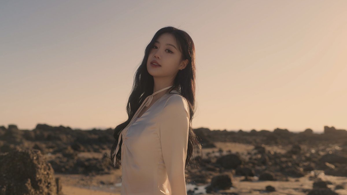 A woman with long dark hair wearing a cream-colored halter top stands on a rocky beach at sunset, looking toward the camera.