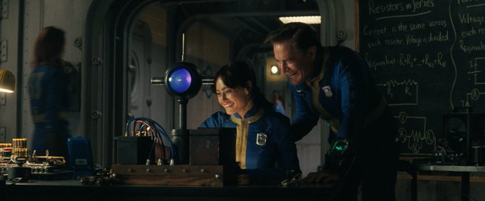 Ella Purnell and Kyle MacLachlan in a first look at the Fallout series coming to Prime Video. They are dressed in bluet and gold suits, and are smiling as they look at something on the desk inside of the Vault they are in.