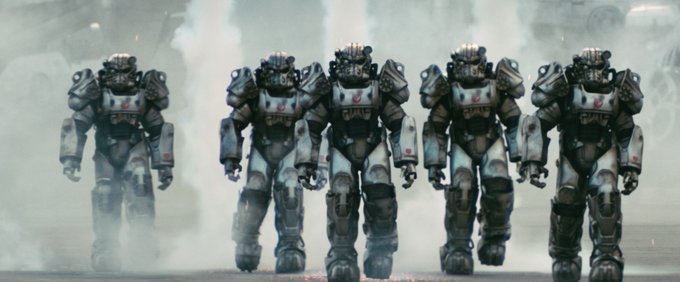 5 Power Armor suits walking together in a first look at the Fallout series coming to Prime Video. They are silver with a red insignia on them. They are very large.