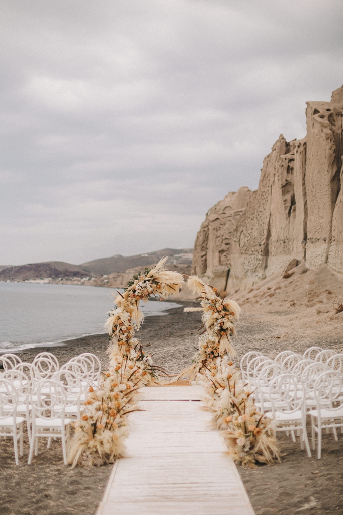 In the magical embrace of Santorini's coastal breeze, a boho-inspired love story dances along the sun-kissed beach. The ceremony, painted against the backdrop of the Aegean's azure canvas, unfolds as an elegant symphony of untamed romance.🧡