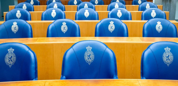 Blauwe zetels in rijen achter de tafels in de plenaire zaal van de Tweede Kamer. 