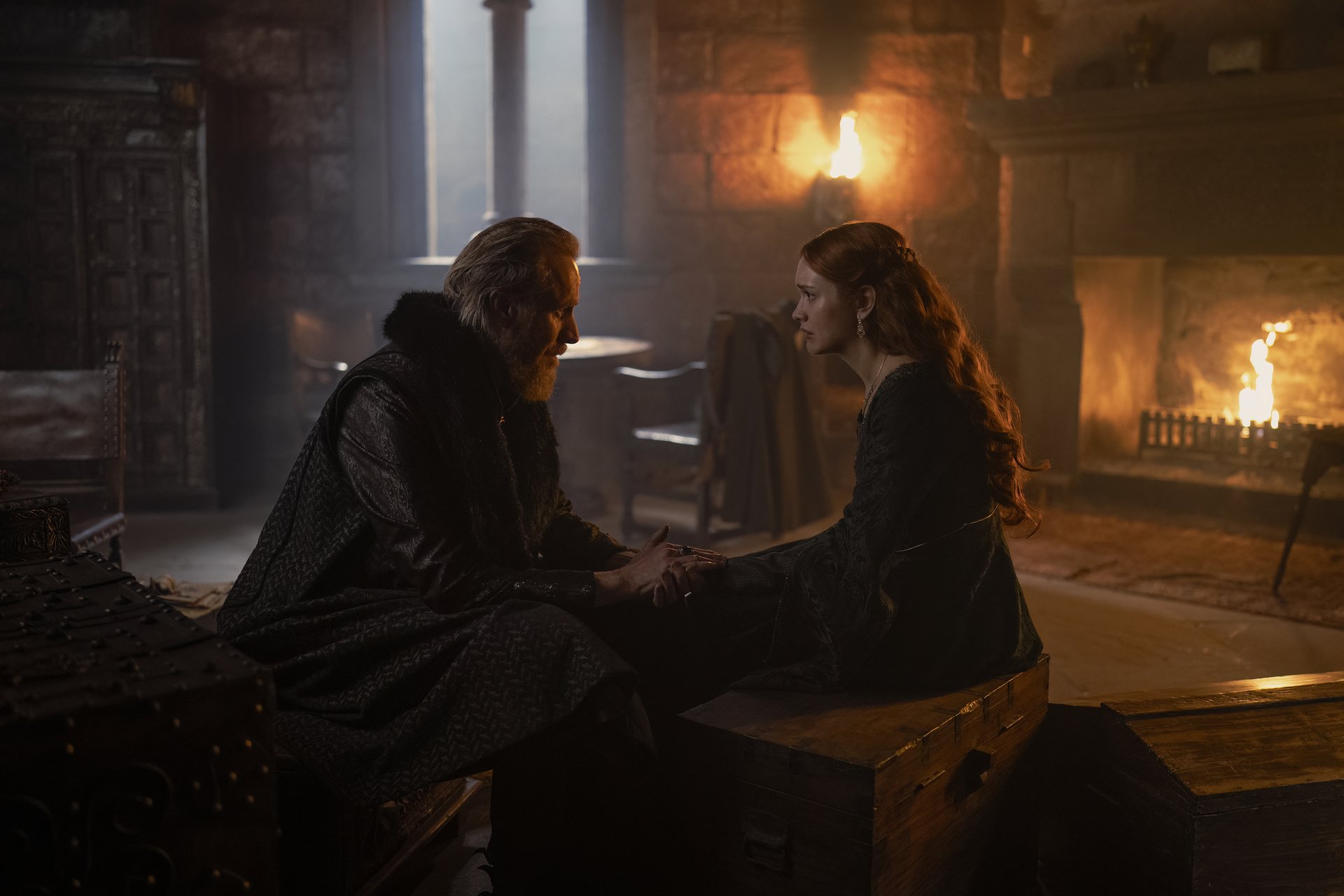 An older man sits with a younger woman and holds her hand. She looks slightly distressed, and is wearing expensive clothing and jewelry. The man is wearing black. They are in an old castle room.
