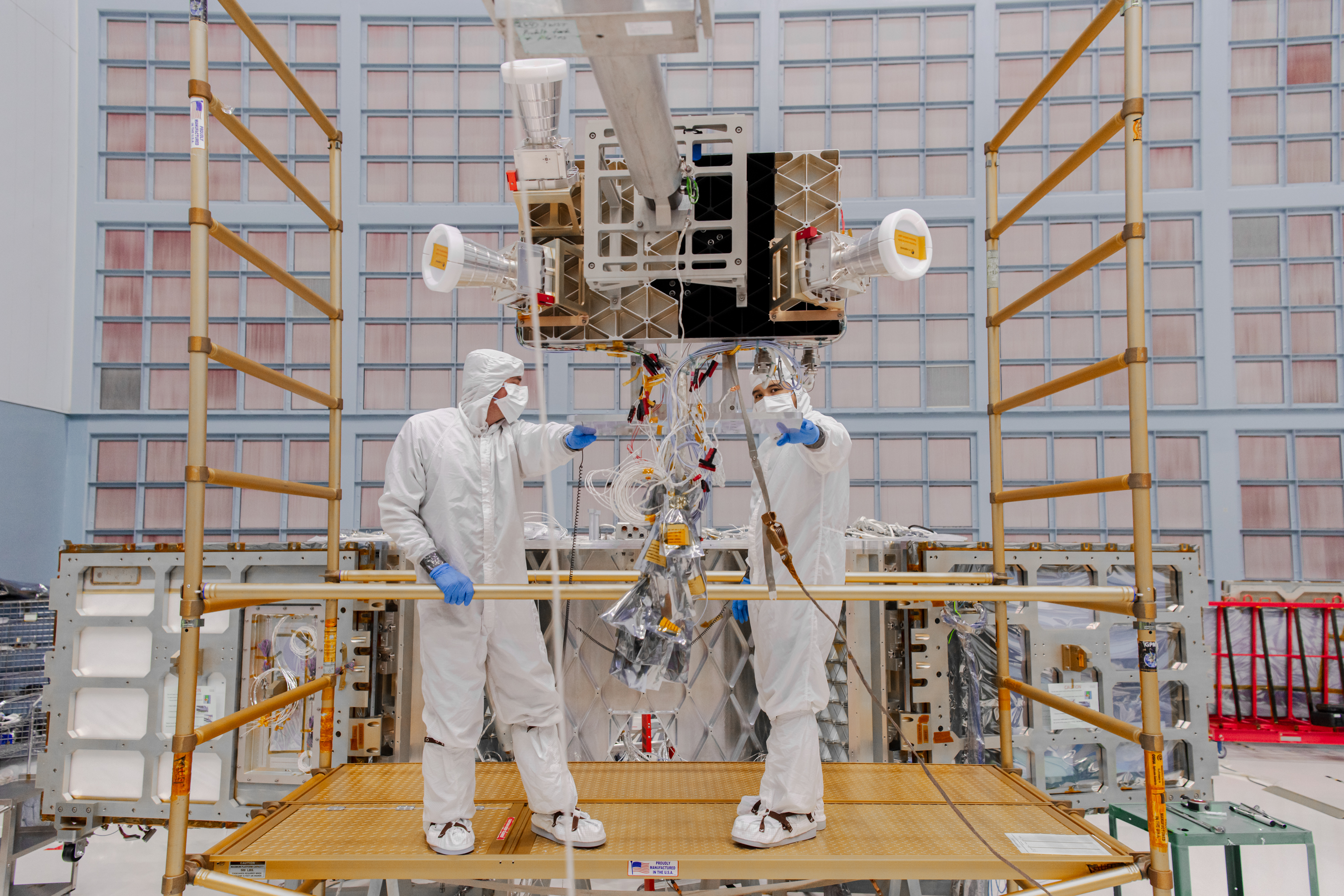 People are in head-to-toe white clean room jumpsuits with blue gloves. They are standing on a gold-colored platform facing forward. In the foreground is a device suspended horizontally from hanging metal rods. At the end of the rods is a large square box, with three white horn-shaped instruments around it. The back wall is covered in a grid of light pink squares, which are air filters. Photo credit: NASA/ Sydney Rohde